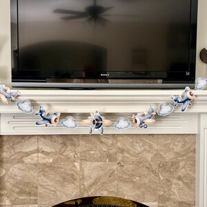 May include: A decorative banner featuring several cartoon airplanes and fluffy white clouds. The airplanes are primarily blue and tan, with propeller details. The banner is strung across a white mantelpiece.
