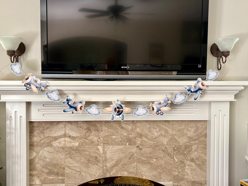 May include: A decorative banner featuring several cartoon airplanes and fluffy white clouds. The airplanes are primarily blue and tan, with propeller details. The banner is strung across a white mantelpiece.