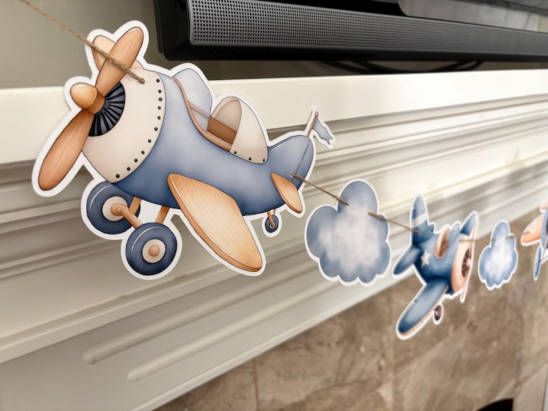 May include: A decorative airplane banner featuring blue and tan watercolor-style airplanes and cloud cutouts. The banner is strung on a natural twine, perfect for a baby shower or nursery decor.