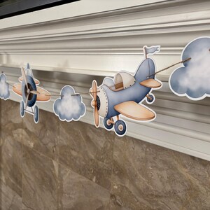May include: A decorative banner featuring paper cutouts of vintage airplanes and clouds. The airplanes are primarily blue and tan, with propeller details. The clouds are white and blue. The banner is strung on a natural twine.