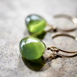 以下が含まれることがあります： 緑色の宝石がついた、金のフープイヤリングのペア。宝石は滑らかで、わずかに輝いています。