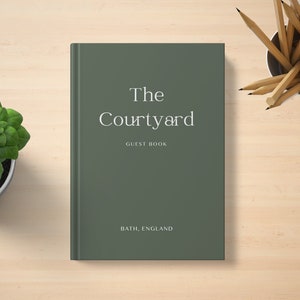 May include: A dark sage green hardback guest book with white text on the front cover that reads The Courtyard GUEST BOOK BATH, ENGLAND. The book is positioned vertically with a potted green plant to the left and a cup of pencils to the right.
