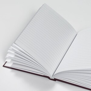 Pu&ograve; includere: Un quaderno aperto con copertina color borgogna e pagine a righe. Le pagine sono vuote e pronte per essere scritte.