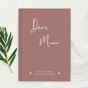 Pu&ograve; includere: Un diario di colore rosa polveroso con il testo "Dear Mum" e "Letters from your daughter" in lettere bianche. Il diario &egrave; su una superficie di legno bianco.