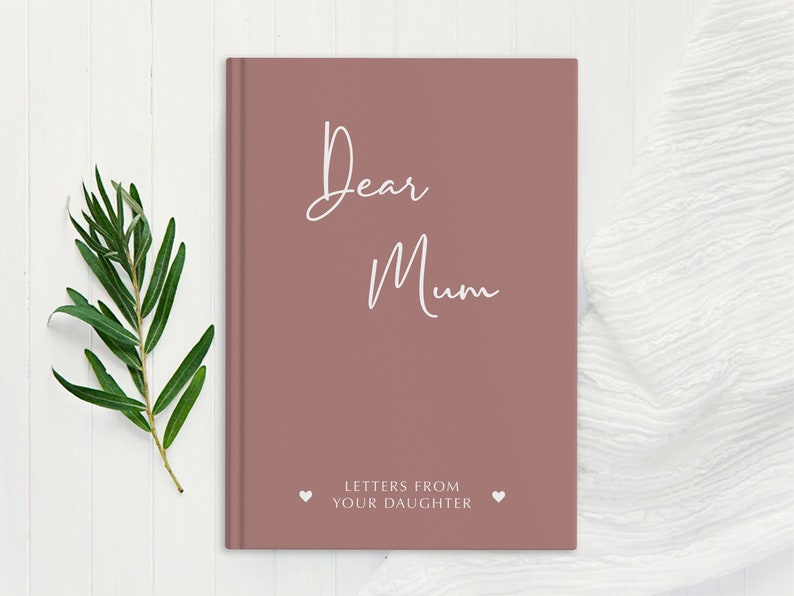 Pu&ograve; includere: Un diario di colore rosa polveroso con il testo "Dear Mum" e "Letters from your daughter" in lettere bianche. Il diario &egrave; su una superficie di legno bianco.
