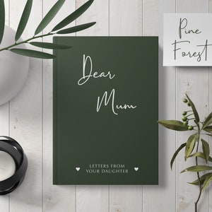 Pu&ograve; includere: Un diario verde scuro con il testo "Dear Mum" in scrittura bianca. Il diario ha le parole "Letters from your daughter" in bianco sotto il titolo. Il diario &egrave; su una superficie di legno bianco con piante verdi e un biglietto bianco con il testo "Pine Forest" su di esso.