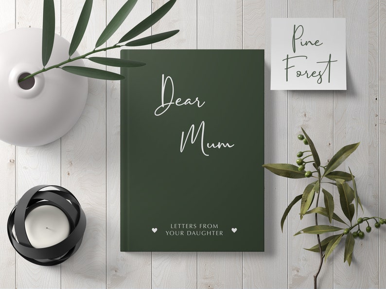 Pu&ograve; includere: Un diario verde scuro con il testo "Dear Mum" in scrittura bianca. Il diario ha le parole "Letters from your daughter" in bianco sotto il titolo. Il diario &egrave; su una superficie di legno bianco con piante verdi e un biglietto bianco con il testo "Pine Forest" su di esso.