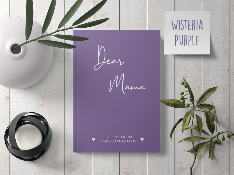 Pu&ograve; includere: Diario viola con il testo "Dear Mama" e "Letters from your daughter" in bianco. Il diario &egrave; su una superficie di legno bianco con un vaso di foglie verdi e un portacandele.