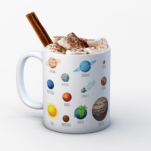 Pu&ograve; includere: Una tazza in ceramica bianca con un'illustrazione colorata dei pianeti del nostro sistema solare, inclusi il sole, la luna e una cometa. La tazza &egrave; piena di cioccolata calda e marshmallow e ha un bastoncino di cannella che sporge dalla parte superiore.