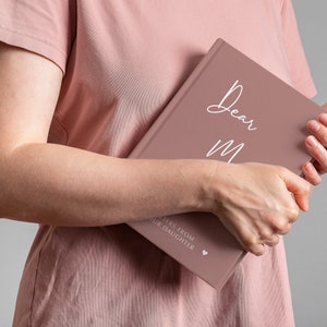 Pu&ograve; includere: Un diario di colore rosa polveroso con il testo "Dear Mom" in scrittura bianca sulla copertina. Il testo "Letters from your daughter" &egrave; stampato in bianco nella parte inferiore della copertina con un piccolo simbolo di cuore.