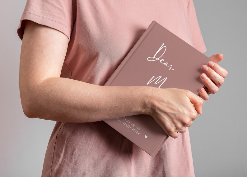 Pu&ograve; includere: Un diario di colore rosa polveroso con il testo "Dear Mom" in scrittura bianca sulla copertina. Il testo "Letters from your daughter" &egrave; stampato in bianco nella parte inferiore della copertina con un piccolo simbolo di cuore.