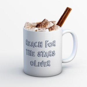 Pu&ograve; includere: Tazza in ceramica bianca con un contorno blu del testo "Reach for the stars Oliver" e riempita di cioccolata calda sormontata da marshmallow e un bastoncino di cannella.