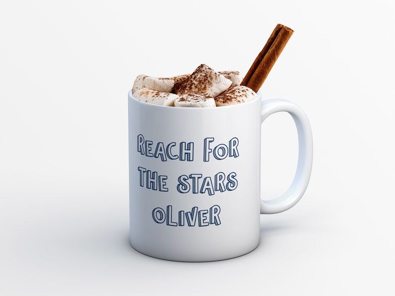 Pu&ograve; includere: Tazza in ceramica bianca con un contorno blu del testo "Reach for the stars Oliver" e riempita di cioccolata calda sormontata da marshmallow e un bastoncino di cannella.