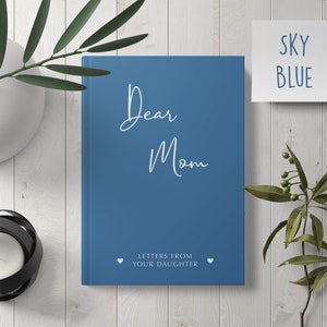 Pu&ograve; includere: Un diario con copertina rigida blu con il testo "Dear Mom" e "Letters from your daughter" sulla copertina. Due cuori bianchi sono su entrambi i lati del testo.