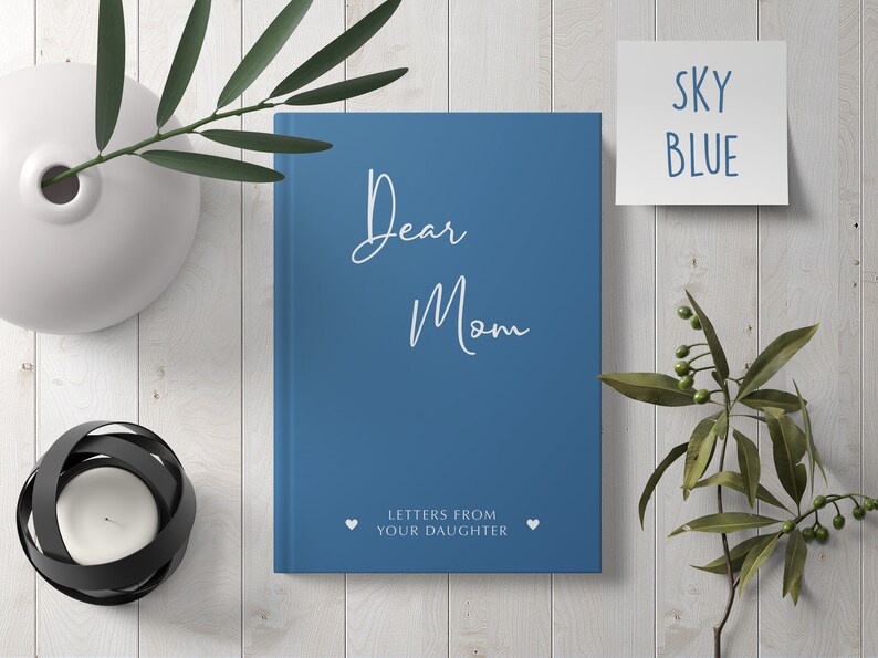 Pu&ograve; includere: Un diario con copertina rigida blu con il testo "Dear Mom" e "Letters from your daughter" sulla copertina. Due cuori bianchi sono su entrambi i lati del testo.