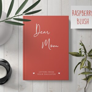 Pu&ograve; includere: Un diario con copertina rigida rossa con il testo "Dear Mom" e "Letters from Your Daughter" in bianco. Il diario &egrave; su una superficie di legno bianco con un vaso di foglie verdi e un portacandele.
