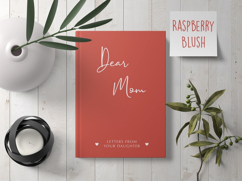 Pu&ograve; includere: Un diario con copertina rigida rossa con il testo "Dear Mom" e "Letters from Your Daughter" in bianco. Il diario &egrave; su una superficie di legno bianco con un vaso di foglie verdi e un portacandele.