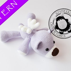 May include: A crocheted purple and white stuffed animal with wings. The animal has black eyes and a brown nose. The animal is lying on its side with its wings spread out. The text "SZYDŁKOWE STWORY CROCHETURES" is visible in a circle in the background.