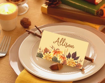 Puede incluir: Un lugar en la mesa con un plato color crema, una tarjeta con el nombre "Allison" y decoraciones florales de otoño. Una bellota y ramitas están en el plato. Una vela encendida y libros en el fondo.