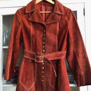 Könnte beinhalten: Eine rostbraune Wildlederjacke mit Gürtel und Knopfleiste. Die Jacke hat weiße Nähte und einen spitzen Kragen.