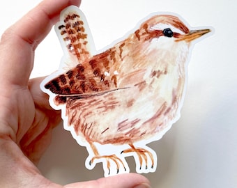 以下が含まれることがあります： 小さな鳥の水彩イラストが描かれたステッカー。鳥は茶色と白の羽、黒い目、黄色のくちばしをしています。ステッカーは白い背景を背景に手で持たれています。鳥の尾羽は濃い茶色の模様で詳細に描かれています。