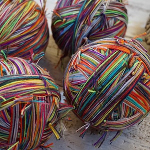 Puede incluir: Primer plano de varias madejas de hilo colorido. El hilo es una mezcla de colores vibrantes, incluyendo rojo, naranja, amarillo, verde, azul y morado. El hilo está enrollado firmemente en madejas, con hebras sueltas visibles.