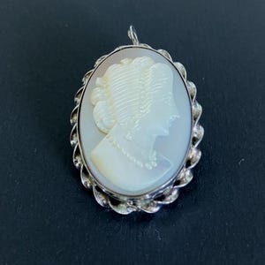 May include: An oval-shaped cameo pendant featuring a carved white profile of a person with an updo hairstyle and a beaded necklace. The cameo is set within a textured, twisted silver-toned metal frame with a small bail at the top for hanging.