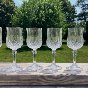 May include: A set of six clear glass goblets featuring a diamond-cut pattern on the lower bowl and a smooth, flared rim. Each glass stands on a slender, multi-faceted stem with a circular base, reflecting light through the textured glass surface.