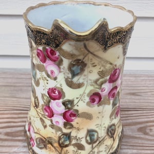 May include: A porcelain pitcher featuring a cream-colored body decorated with hand-painted pink and dark red roses, green foliage, and gold filigree accents. The rim and base are scalloped with a black border and gold trim detailing.