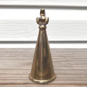 May include: A polished brass bell with a smooth, conical body and a simple, rounded handle at the top. The metal surface reflects light, showing a warm, golden-yellow tone throughout the structure. The bell features a clean, minimalist design.