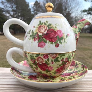 May include: A white porcelain tea-for-one set featuring a teapot stacked on a teacup and saucer. The teapot has a gold knob, while the cup and saucer display a vibrant pattern of red, pink, and cream roses on a light green background with gold trim.