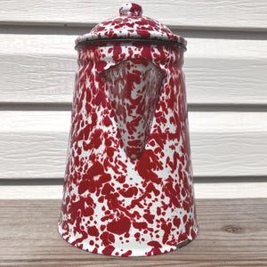 May include: A tall, cylindrical enamel coffee pot featuring a white base with a red speckled, marbled pattern. The pot includes a matching lid with a small knob handle and a triangular spout integrated into the upper body of the vessel.