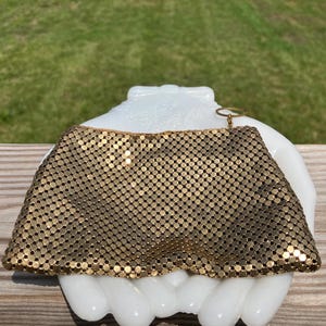May include: A gold-toned metal mesh pouch featuring a fine, interlocking link construction. The rectangular accessory has a smooth top edge and includes a small, circular metal ring attached to the upper right corner for hanging or carrying purposes.