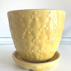 May include: A yellow ceramic flower pot with a matching saucer. The pot has a textured surface with a diamond pattern and small, raised flower designs. The pot is sitting on a matching saucer.