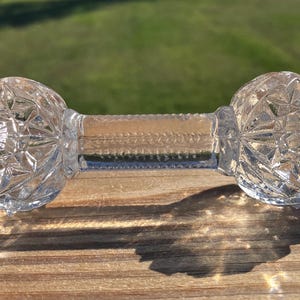 May include: Clear glass dumbbell-shaped object with faceted, round ends and a textured center. The object is resting on a wooden surface, casting a shadow. The background is blurred green.