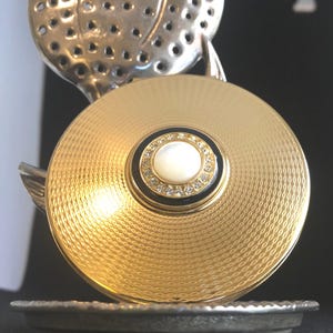 May include: A gold-toned compact with a textured surface and a central design featuring a pearl-like center surrounded by rhinestones. A silver-colored, perforated floral design is visible in the background. The compact sits on a silver-colored tray.