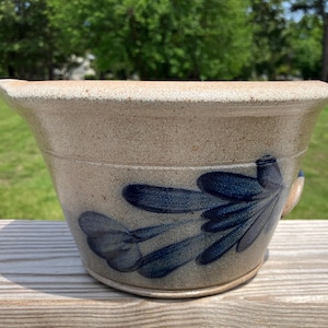 May include: A beige stoneware mixing bowl with a wide rim and a small pouring spout. The exterior features a hand-painted dark blue floral design with elongated petals. The surface has a speckled, textured finish consistent with fired clay pottery.