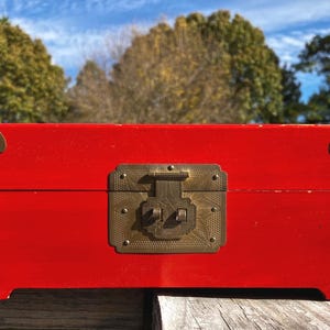 May include: A rectangular, red wooden box with brass corner accents and a central latch. The box has decorative brass hardware and is set against a backdrop of trees and a blue sky. The box is approximately 12 inches long.