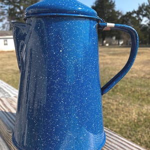 May include: A blue enamelware coffee pot with white speckles. The pot has a lid, a handle, and a spout. It is sitting on a wooden surface. The background is a sunny outdoor scene.