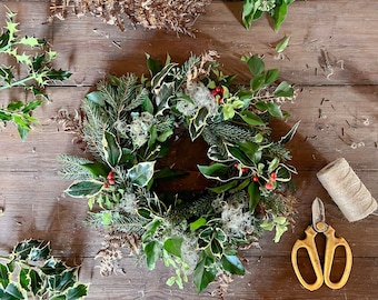 May include: A handmade Christmas wreath with green and white variegated holly leaves, red berries, and sprigs of evergreen. The wreath is surrounded by additional greenery, scissors, and twine on a wooden surface.