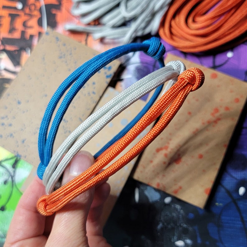 May include: Three paracord bracelets in blue, white, and orange, tied together in a knot. The bracelets are made of woven paracord.