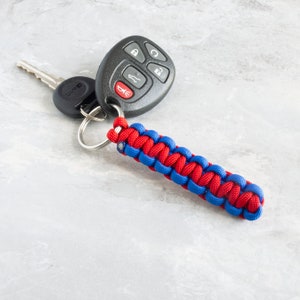 May include: A black GMC car key and remote fob attached to a metal split ring with a braided paracord keychain. The keychain features a repeating pattern of vibrant red and bright blue nylon cord woven into a thick, textured cylindrical shape.