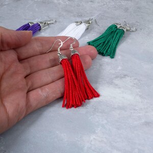 May include: Red tassel earrings with silver-tone metal accents. Additional earrings in white, green, and purple are visible in the background. The earrings are displayed on a light gray surface.
