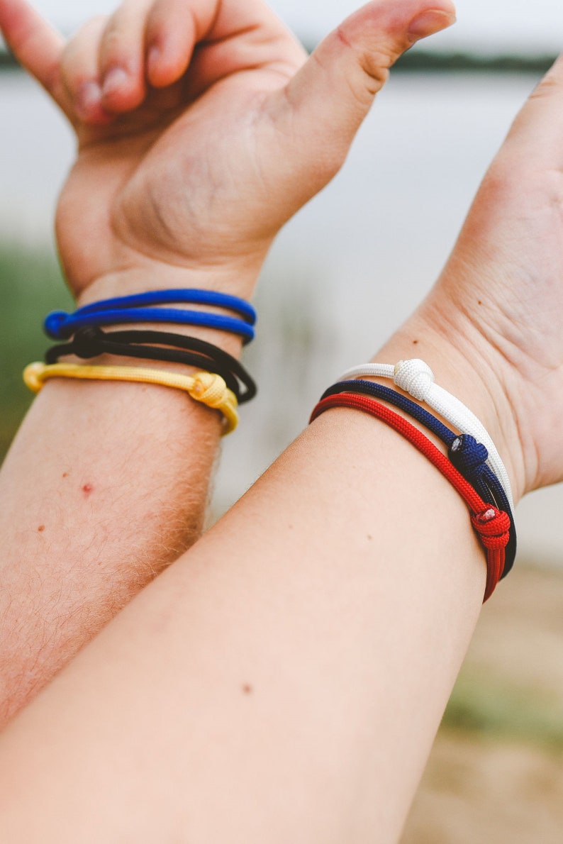 close up of 2 arms, one wearing blue, black and yellow bracelets, and the other wearing red, white and blue bracelets