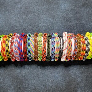 May include: A collection of colorful paracord bracelets displayed on a white cylindrical object. The bracelets feature various patterns and colors, including green, yellow, red, blue, and black. Each bracelet is approximately 0.5 inches wide.