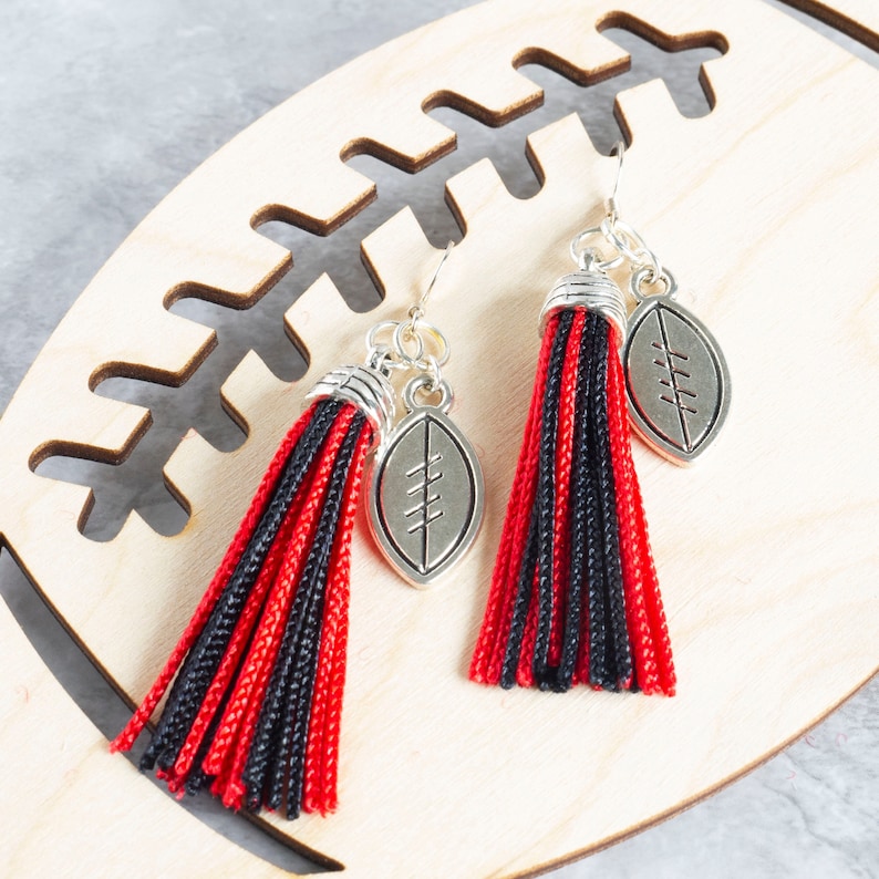 Red and black tassel earrings with silver football charms, laying on a wooden cut out of a football