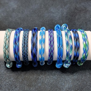 May include: A collection of eight blue paracord bracelets, each with a unique pattern and shade of blue. The bracelets are displayed on a white cylindrical surface, showcasing the variety of designs and colors.