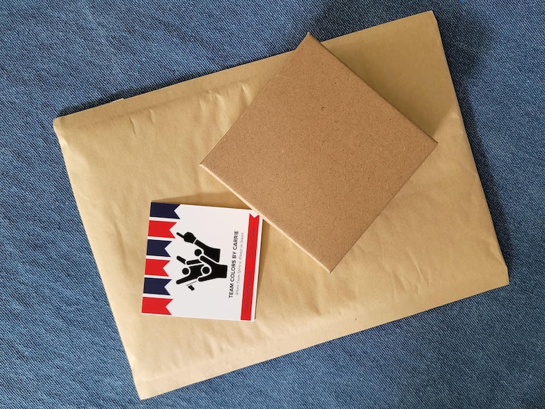 May include: A kraft paper envelope and a brown cardboard box rest on a blue denim surface. A small card with red, white, and blue stripes and the text "TEAM COLORS BY CARRE" is also present. The items are arranged for shipping.