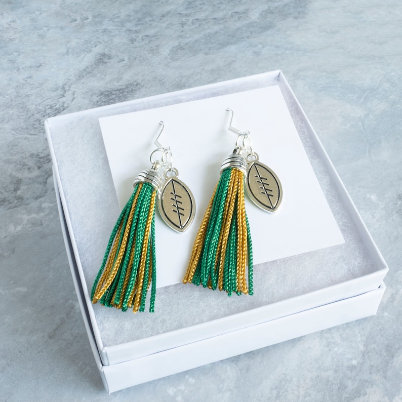 May include: A pair of green and gold tassel earrings with a silver football charm. The earrings are displayed in a white gift box.