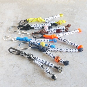 May include: A set of nine colorful keychains with different sports themes. Each keychain features a small ball charm and beads with the words "MOM", "DAD", "SOCCER", "BASKETBALL", "VOLLEYBALL", "FOOTBALL", "BASEBALL", "SOFTBALL", and "TENNIS".